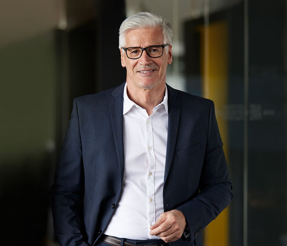 Mark Ram standing in a office hallway wearing a white button up with dark blue jacket and dark rimmed glasses