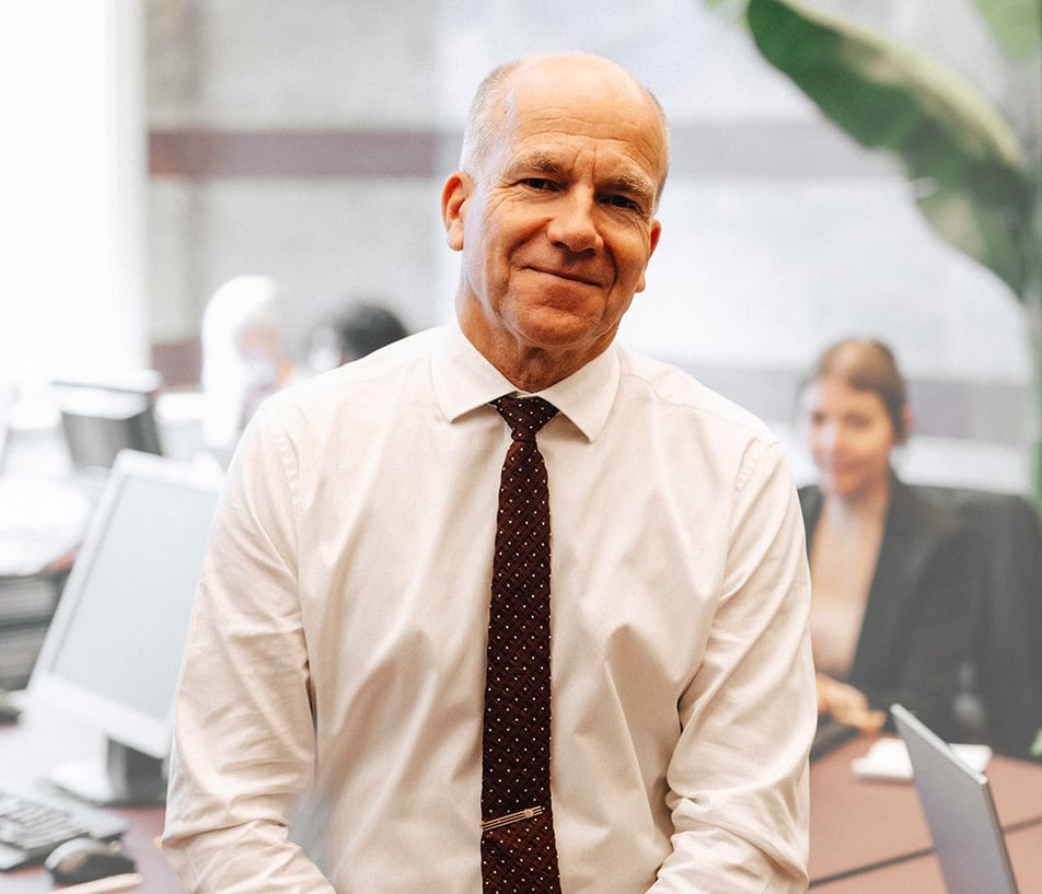 Brian Grott wearing a white button up with a red tie with white dots, softly smiling and leaning against a desk