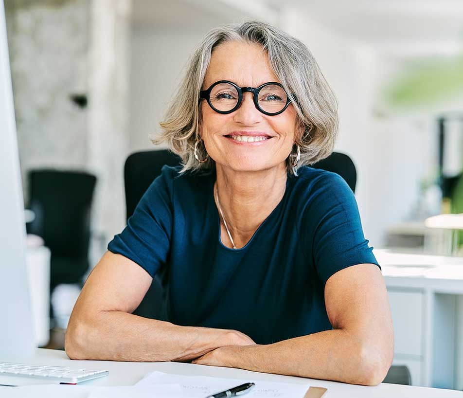 Susan Farina wearing a dark blue top and dark rimmed circular glasses brightly smiling while sitting at a desk with a pen and paperwork in front of her